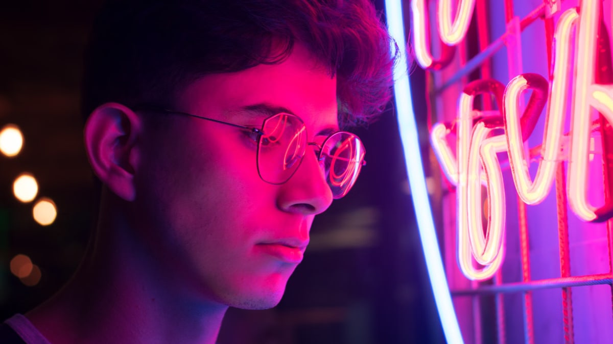 A young man with glasses illuminated by vibrant neon lights in an urban setting.